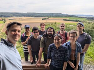Group photo, from left to right: Vasily, Subhadip, Omar, Daniel, Soham, Pritha, Lukas, Miriam, Ricardo. In the background one can see fields and a forest on a sunny day.