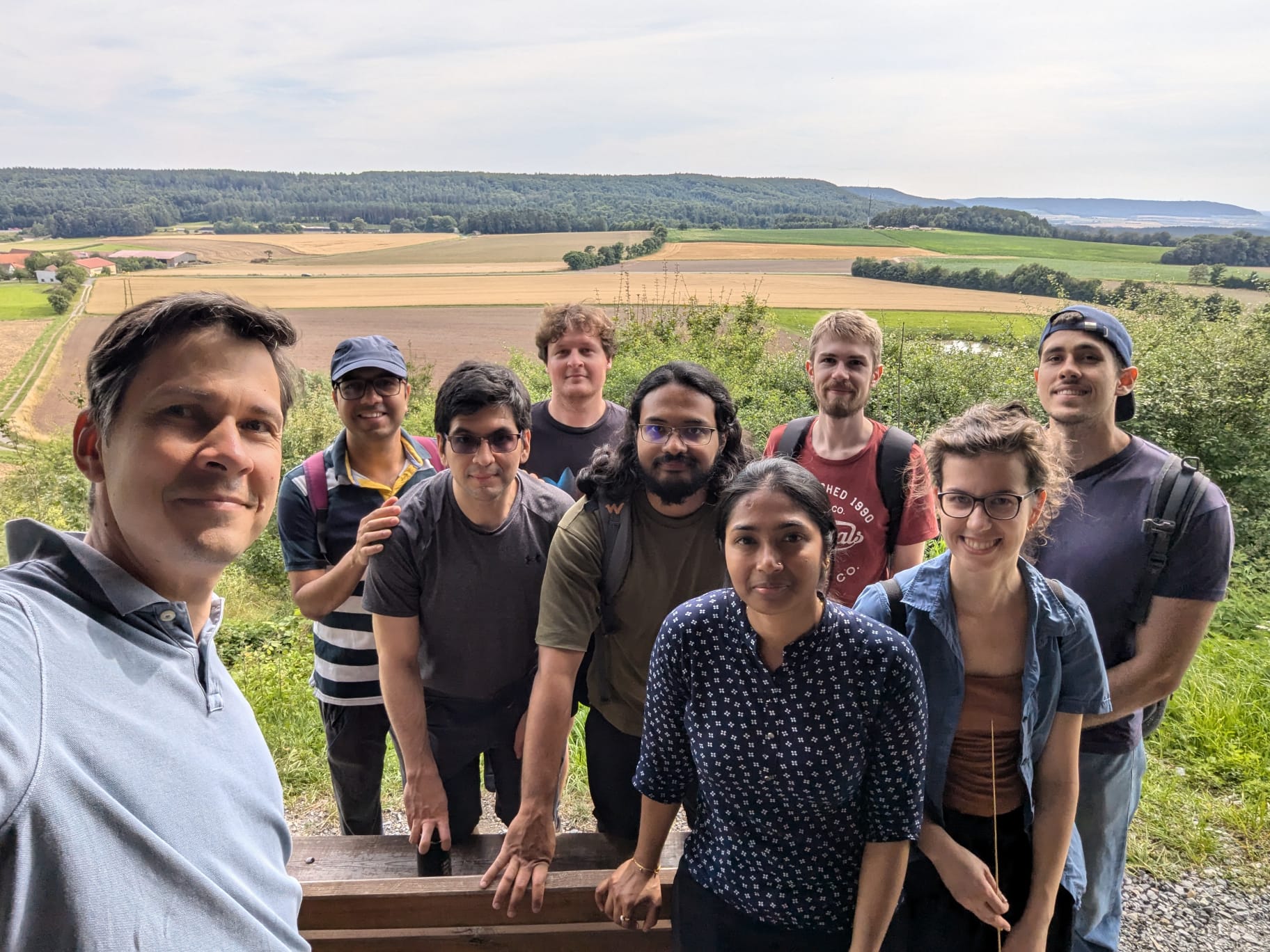 Group photo, from left to right: Vasily, Subhadip, Omar, Daniel, Soham, Pritha, Lukas, Miriam, Ricardo. In the background one can see fields and a forest on a sunny day.