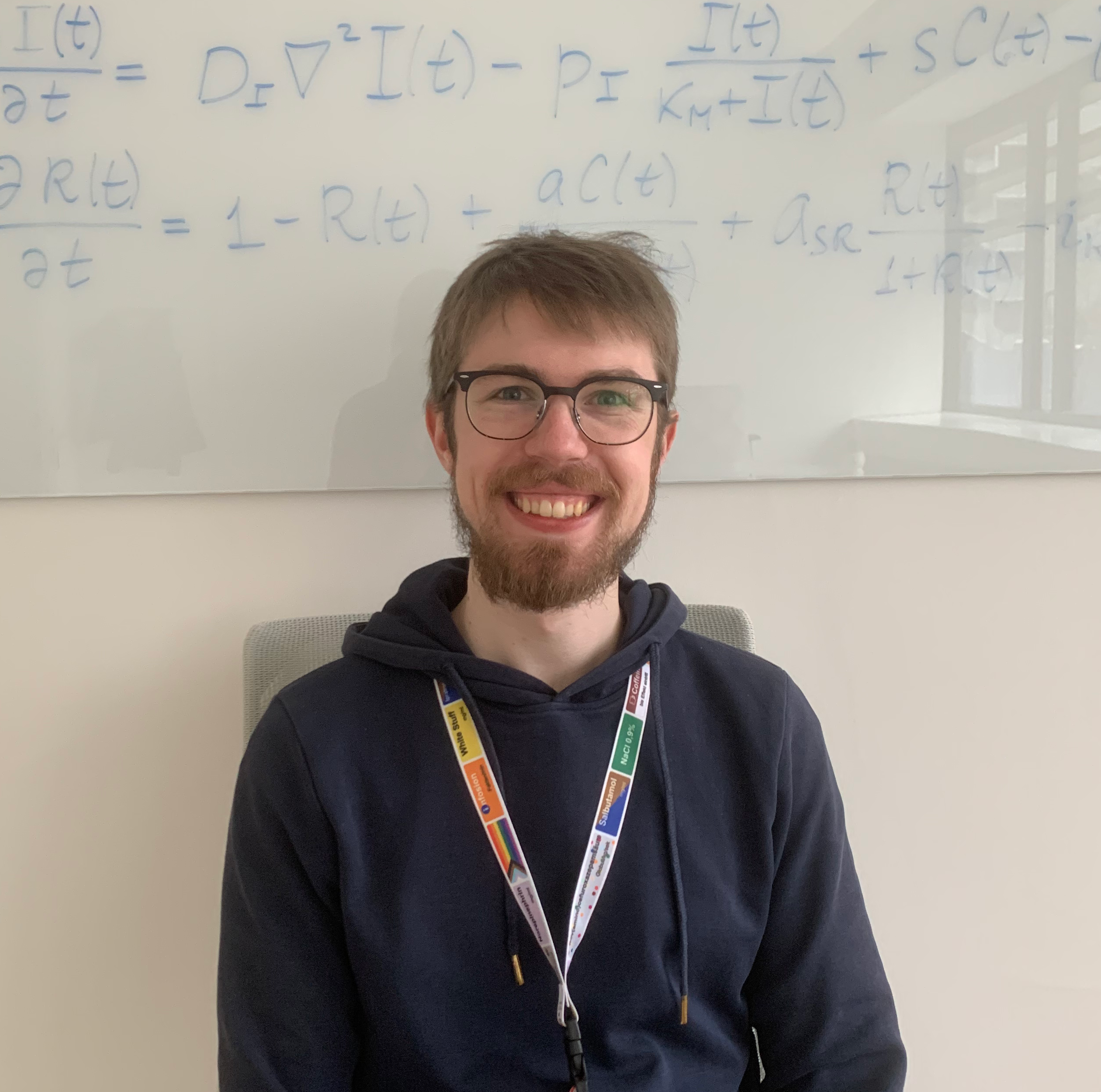 Portrait of Lukas Pöschl. Lukas sits in front of a white board with mathematical equations. He has short brown hair and a beard, wears glasses and smiles towards the camera