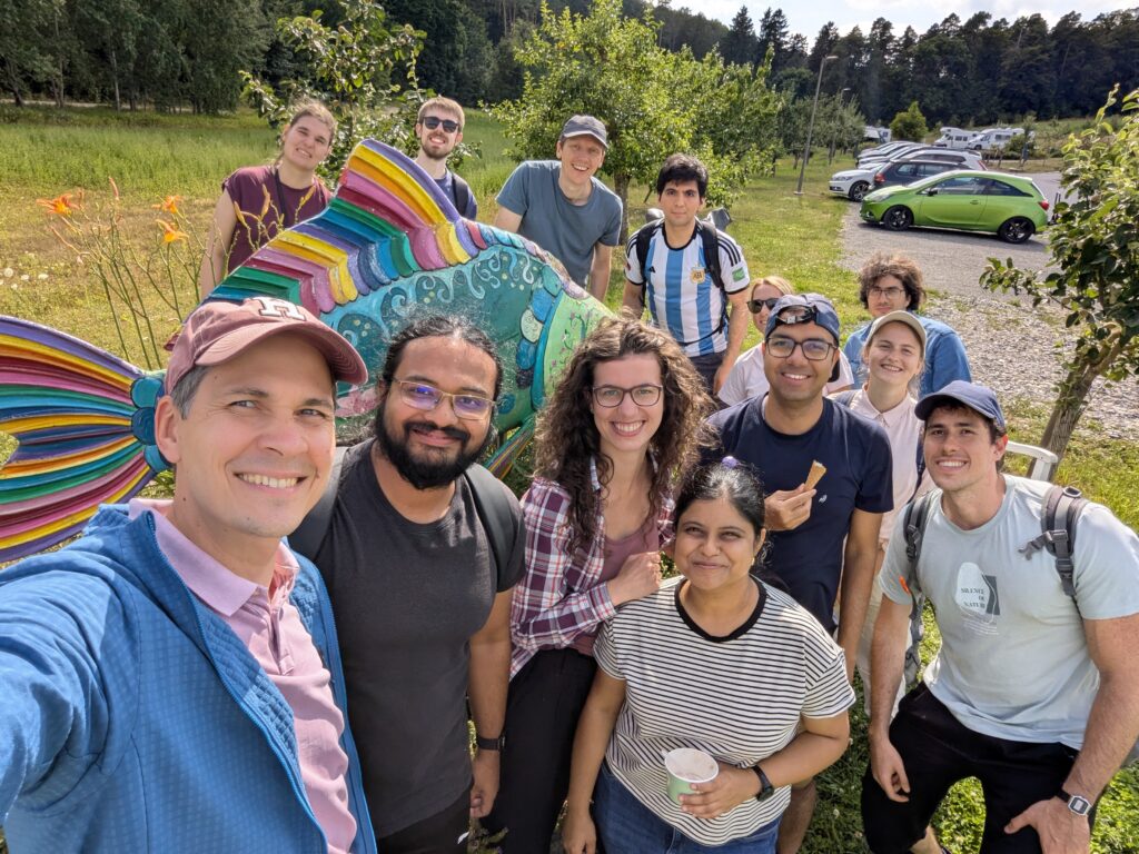 Selfie group picture of the research group. The people are standing outside around a statue of a colourful fish on a sunny day. Two people are holding ice cream.