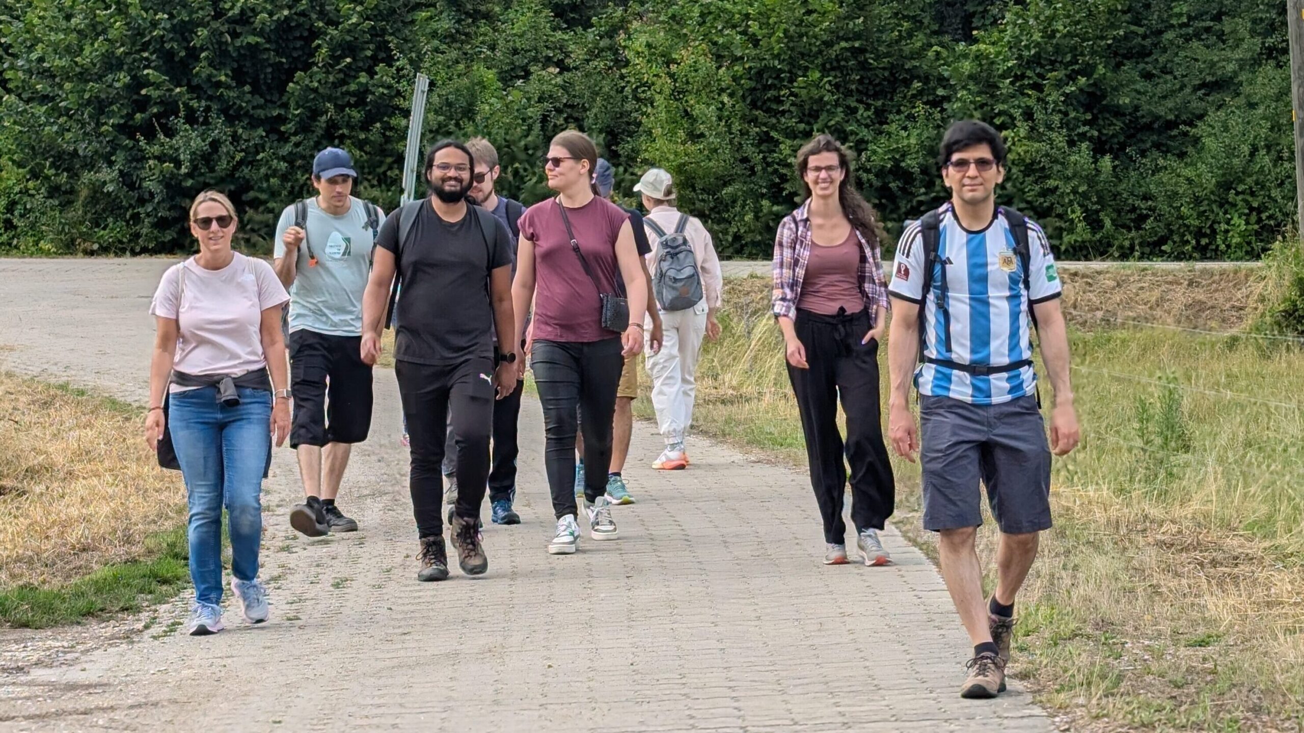 Multiple people walking on a paved way. It is sunny, as some people are waring sun glasses, in the background there are large trees and bushed