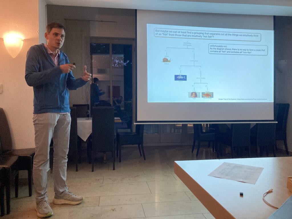 In a seminar room, Vasily wears a blue jacket and stands next to a projected powerpoint presentation slide. He gestures as he is presenting something.