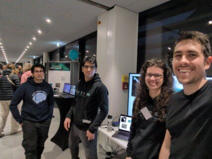 Group picture of Ricardo, Miriam, Omar and Mathar, standing in a hallway in front of a monitor. They are all wearing black pullovers with the MPZPM logo on the front.
