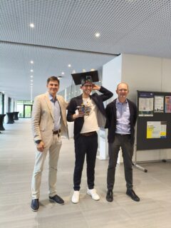 Vasily Zaburdaev, Tim Klingberg and Lennart Hilbert stand in a hallway of MPZPM and smile into the camera. They are wearing suits, Tim is wearing a doctoral hat on his head and holds another smaller doctoral hat in his hand.