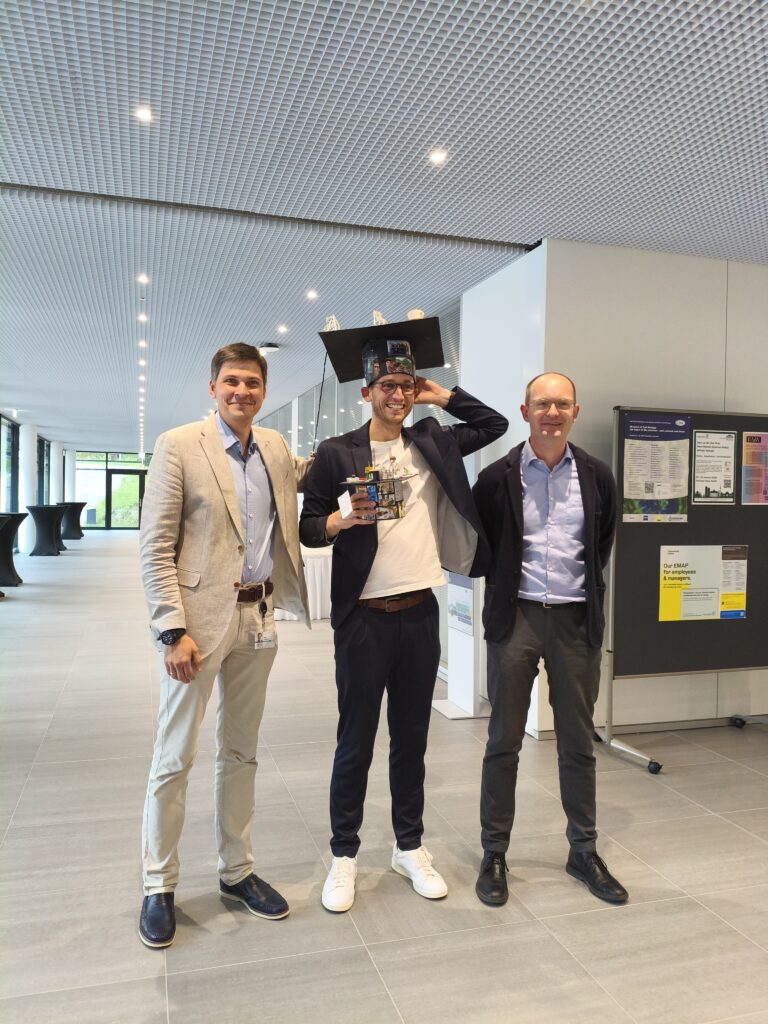 Vasily Zaburdaev, Tim Klingberg and Lennart Hilbert stand in a hallway of MPZPM and smile into the camera. They are wearing suits, Tim is wearing a doctoral hat on his head and holds another smaller doctoral hat in his hand.